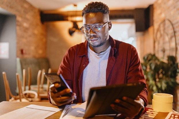  A male coffee shop owner sits in his shop. Before him are papers. He is holding a computer tablet in his left hand a smartphone in his right hand. He is comparing financial data.