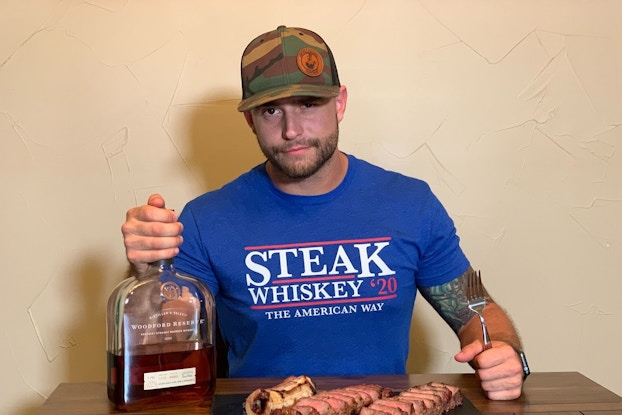 A man wears a hat and sits a table with grilled meat and a liquor bottle beside him.