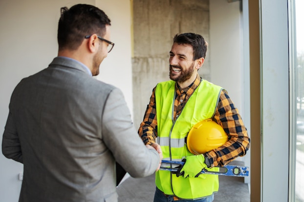  General contractor with safety vest and hardhat smiles and shakes hand of man in a business suit.