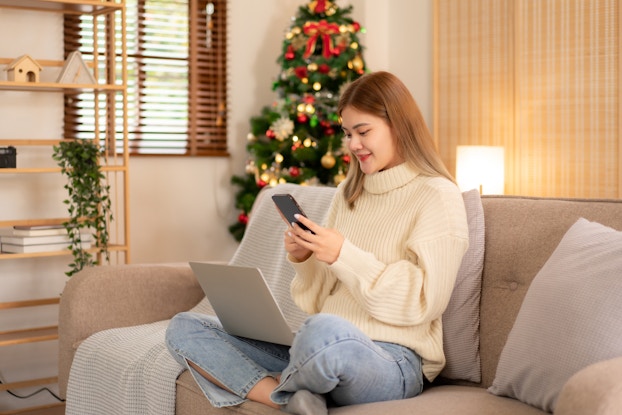  Woman sitting on her sofa in her living room smiling at her phone and laptop while shopping.