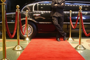  Limo driver in a suit leaning against a limo in front of a red carpet. 