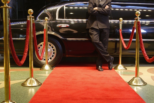 Limo driver in a suit leaning against a limo in front of a red carpet.