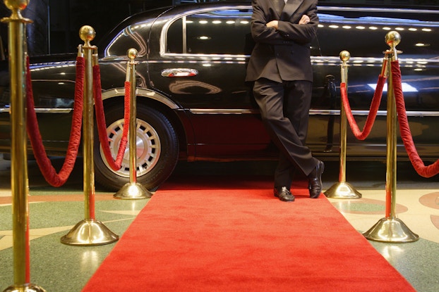 Limo driver in a suit leaning against a limo in front of a red carpet.