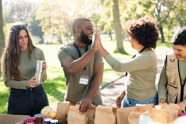 A group of four volunteers are shown in front of a table. Two individuals are high-fiving each other. Before them on a table is a group of bagged sandwiches and snacks they've prepared to hand out.