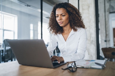 A businesswoman is seated at a table in a modern office. She has a laptop open before her and she is typing on it.