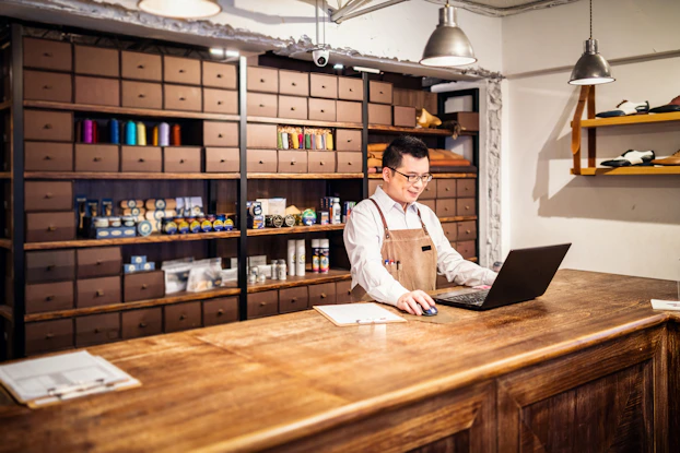 A shop owner works at his laptop in front of a wall of sewing and shoe care products.