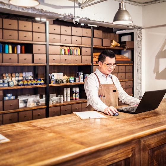 A shop owner works at his laptop in front of a wall of sewing and shoe care products.