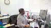 A woman sits in front of a desktop computer. She is holding payroll checks in her hand and looking at the computer monitor.