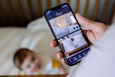A close-up of a hand holding a smartphone showing the Harbor baby monitor app's gallery view. The screen is split between a video of an empty bed and an overhead shot of a crib with a baby in it. In the background beyond the phone, out of focus, is the same crib and baby seen on the phone screen.