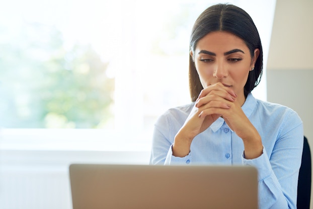 Serious young businesswoman with a worried expression sitting reading information on her laptop with clasped hands.