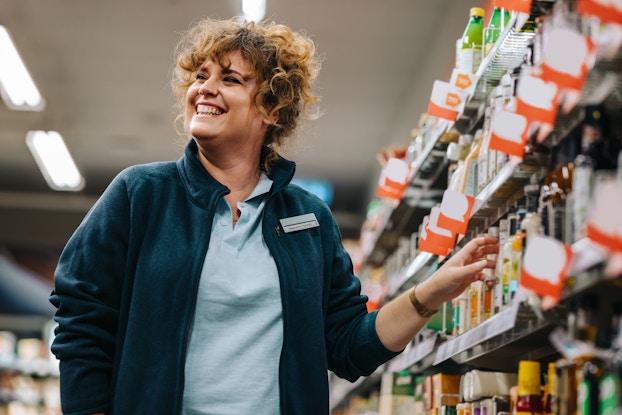  Confident grocery story employee standing by the shelves and smiling in supermarket.