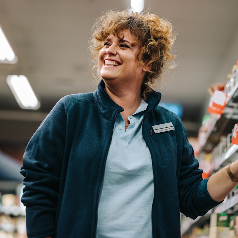  Confident grocery story employee standing by the shelves and smiling in supermarket. 