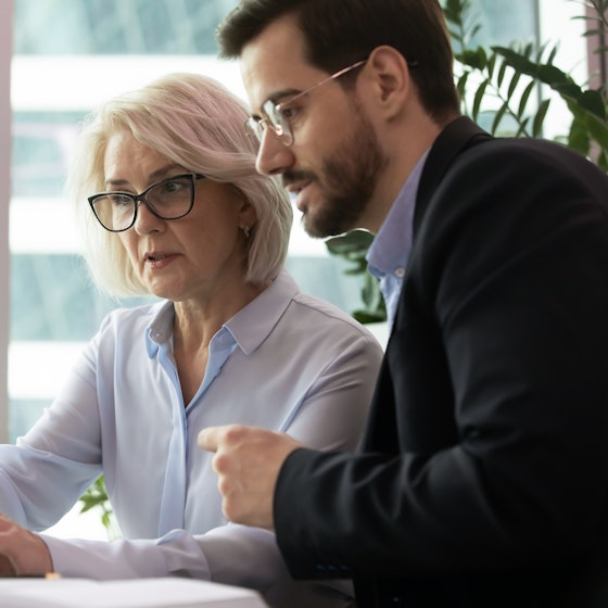 Businesswoman communicates with younger businessman carefully look at laptop screen.