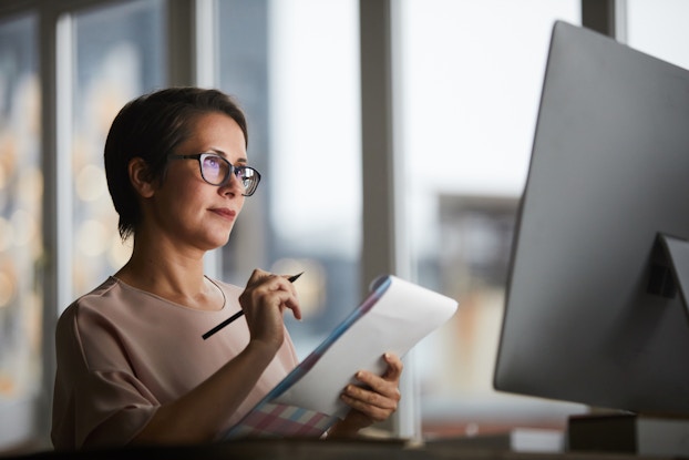  Woman looks at desktop computer screen and takes notes on a notepad.