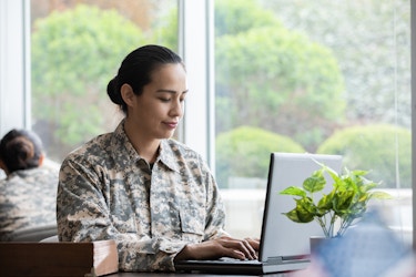  Woman wears military uniform and works on laptop in an office. 