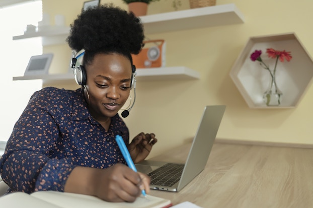 Smiling woman employee wears a headset and takes notes in a notebook while using her laptop computer.