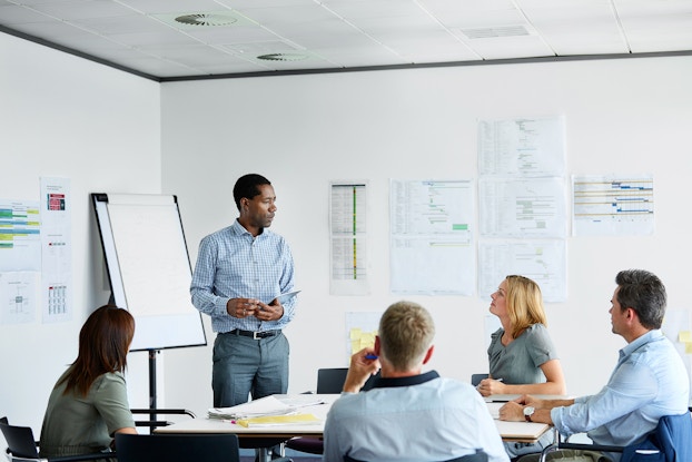 Man stands in meeting room with charts on the wall, presenting a strategy to four colleagues.