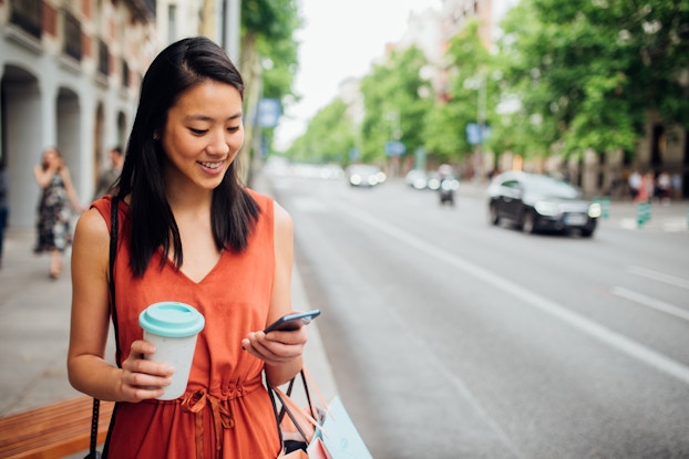  A woman holds a travel coffee mug while she checks her phone on a city sidewalk.