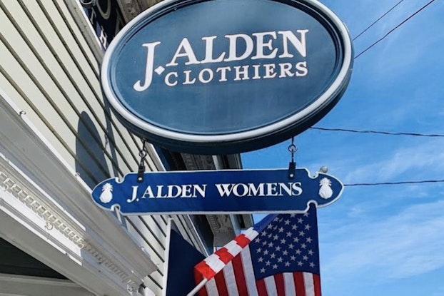  A storefront sign hangs outside a store, American flag waving behind it.