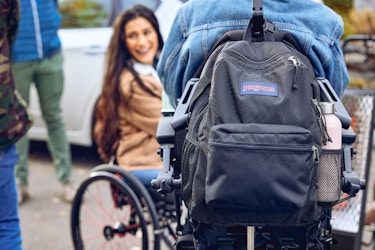  Back image of a person wearing an adaptive backpack by JanSport, with a girl in a wheelchair in the background. 