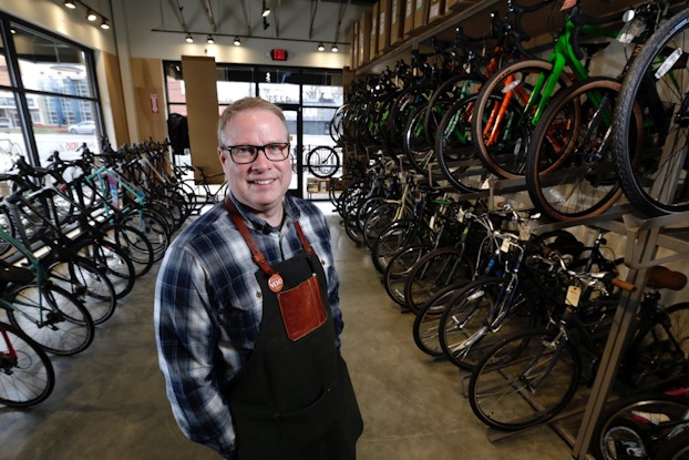 A smiling man wearing glasses, a plaid shirt and an apron stands in a bike shop with rows of bikes behind him.