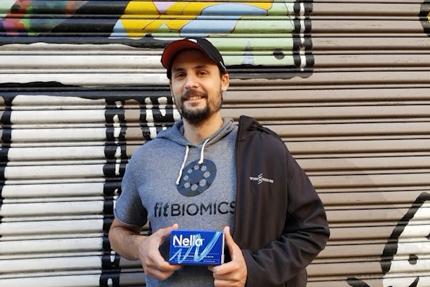 A shot of Dr. Jonathan Scheiman, Co-founder and CEO of FitBiomics, standing in front of a mural and holding a small blue box of FitBiomics' Nella probiotics capsules. Jonathan is a white man with a beard. He's wearing a black-and-red baseball cap and a hooded gray T-shirt with the FitBiomics logo in black on the front. Draped over one shoulder is a black hoodie with FitBiomics's logo on one side.