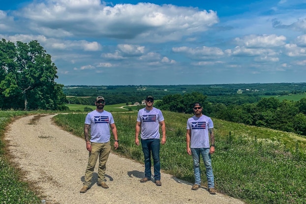 Three men wear matching t-shirts, standing on a country dirt road in front of a green landscape and blue sky