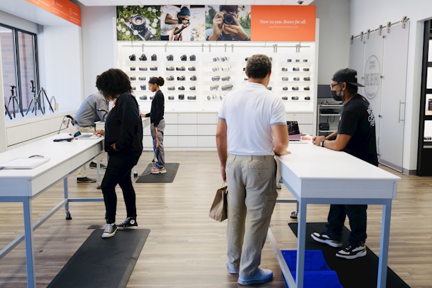 Five people browse KEH Camera's store in Atlanta, Georgia. The store is white-walled, with narrow white tables standing at regular intervals throughout the room. Refurbished cameras are mounted on the back wall and stand on tripods on a windowsill on the left side of the room. Customers stand at the tables, using laptops or waiting for assistance.