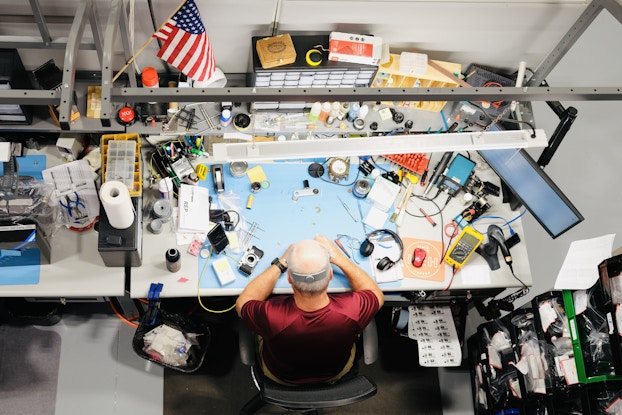 A view from above of a worktable at KEH Camera, where a technician inspects camera parts and other pieces of tech. The worktable is large and scattered with camera parts and handheld tools. A computer monitor, an American, flag, and a couple of storage organizers sit further back on the table. A camera with its lens removed sits near the technician, whose head obscures what his hands are working on. The technician is a balding man wearing an orange T-shirt. He sits in a wheeled office chair as he works.