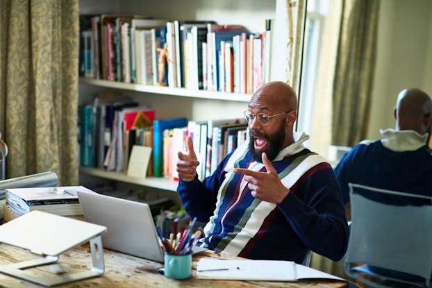 An entrepreneur who is seated at a desk before a laptop and is the middle of a video conference responds animatedly to something he has heard.