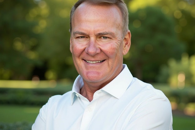 A headshot of Kelly Roddy, CEO of WoWorks. Kelly is a middle-aged white man with blond hair. He wears a white collared shirt and he is smiling for the camera. He is standing outside; in the background, out of focus, is a large grassy area bordered by trees.