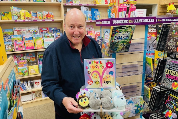 Kenny Sarfin, Owner of Books & Greetings, stands next to a display for Beanie Bouncers. Kenny is an older man wearing a dark blue fleece jacket and smiling for the camera. The display comes up to Kenny's elbows in height. It has two tiers filled with little round plush toys, including black-and-yellow bees, gray koalas, and pale blue axolotls. On top of the display is a square sign showing cartoons of the Bouncers bouncing around between the phrases "We Bounce" and "I Bounce." The price, $5.99, is written in black at the lower-right corner of the sign.