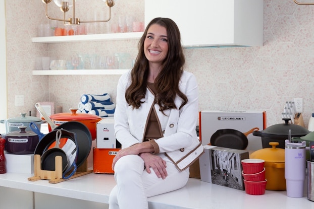 Brittany Valentine, one of the Founders of Kitchen Switchen, sits on a kitchen counter surrounded by refurbished and secondhand cookware. Brittany is a young woman with long brown hair. She wears an olive green shirt, a white jacket, and white pants. The cookware around her includes a set of skillets in a wooden rack, a few brightly colored crockpots, and a small stack of red ceramic ramekins.