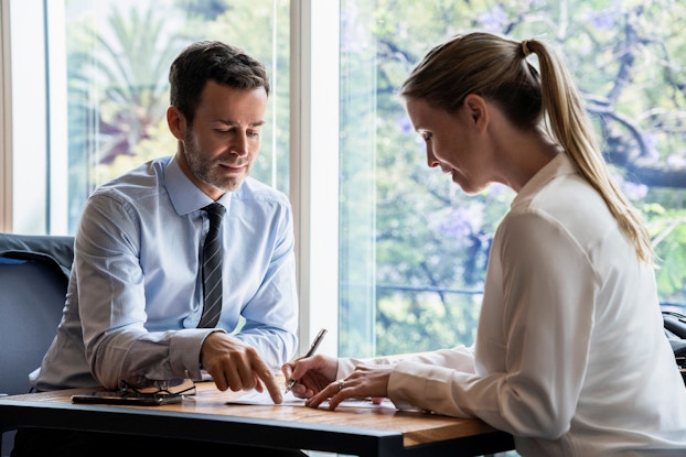 A man and a woman are seated at a table. The man, a lawyer, points at a document to show the woman, a business owner, where to sign. She is signing legal papers to form a limited liability company.