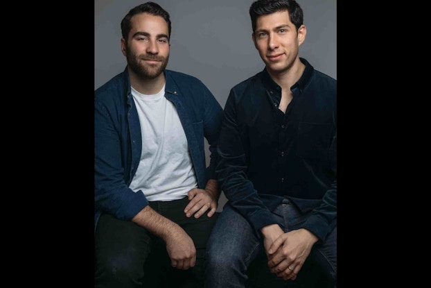 Michael Weider (left) and Greg Davidson, the Co-founders of Lalo, sitting side-by-side in front of a gray background. Michael is dark-haired with a beard, and he wears a dark blue button-up shirt over a white T-shirt and dark pants. Greg is dark-haired and wearing a dark blue button-up shirt and jeans.
