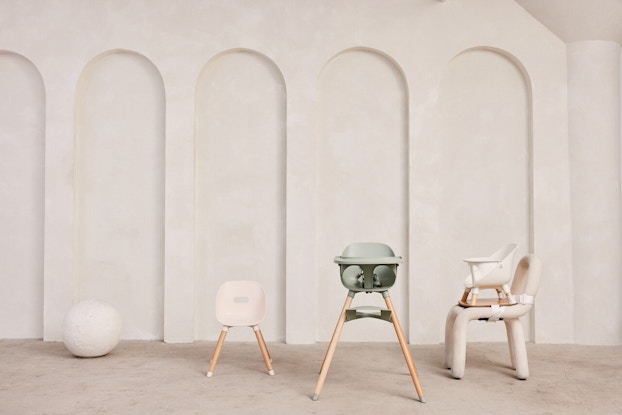 Three of Lalo's baby chairs, standing in a neutral room with white walls. From left to right, they are the Play Chair (a child-sized white chair, for use at a play table), the Chair (a three-in-one highchair with tray), and the Booster (a white booster seat standing on and strapped to a regular-sized chair).