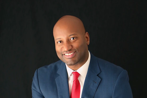 A photo of Lonnie McGowen, a Franchisee of IMAGE Studios, smiling and seated against a black background. Lonnie is a Black man wearing a navy blue suit and red tie.