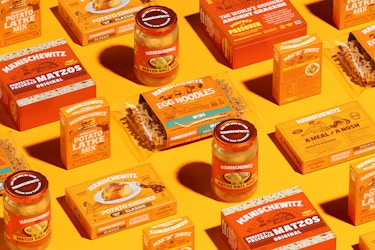 Manischewitz brand packaged foods are arranged in a pattern against a mustard yellow background. The foods include potato latke mix, boxed potato knishes, jarred matzo ball soup, bagged egg noodles, and boxes of matzos labeled "Perfect for Passover."