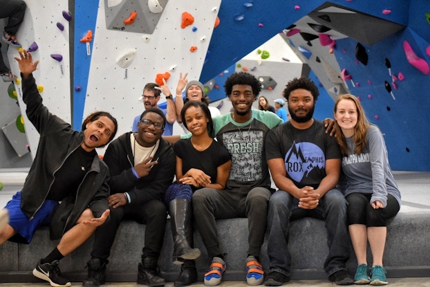 A jovial group of smiling people sit in a rock climbing room.