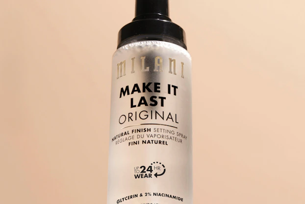 A bottle of Milani's Make It Last setting spray. The bottle is frosted white glass with a black nozzle on top.