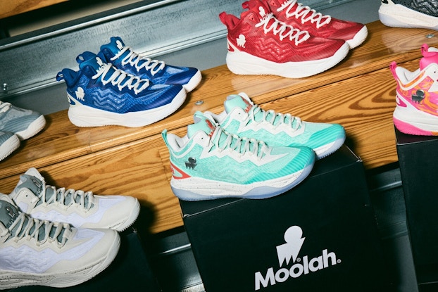 Two display rows of Moolah Kicks sneakers on display -- the back row of gray, blue, and red sneakers are on a wooden bench and the front of pale blue, mint green, and pink sneakers are sitting on individual black blocks.