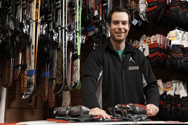 An owner of a ski shop smiles directly at the camera. Before him on a table is a pair of skis.