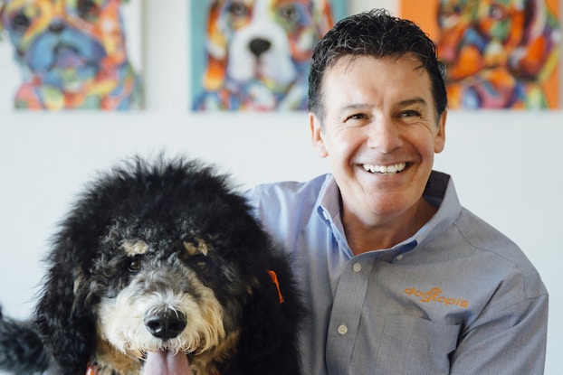  A headshot of Neil Gill, CEO of Dogtopia. He is sitting to the right of a large black-and-white dog wearing an orange bandanna and red collar.
