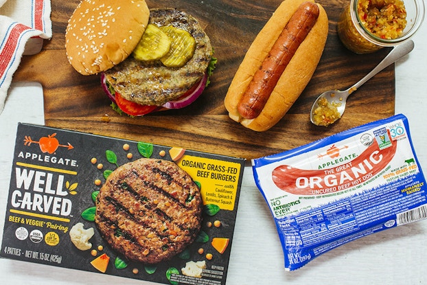  Packages of Applegate hot dogs and hamburgers displayed on top of a board with the cooked food.