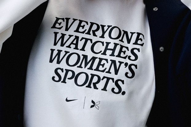 A close-up of a white T-shirt that reads "EVERYONE WATCHES WOMEN'S SPORTS." in all-caps black type. The Nike swoosh and the Togethxr logo (a half-black half-white stylized X) are printed beneath this words.