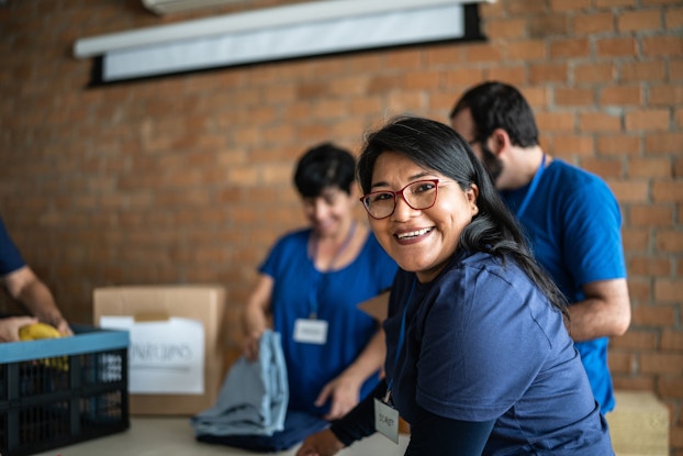  A woman volunteer working in a community charity donation center smiles at the viewer. Behind her are other volunteers who are folding clothes.