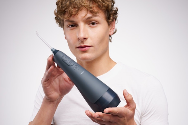 A teen boy holds up one of Ordo's sonic water flossers. The flosser has a gray base that narrows toward the top and a thin white head that sprays water. The teenager has curly blond hair and wears a white T-shirt, and he looks at the viewer with a neutral expression.