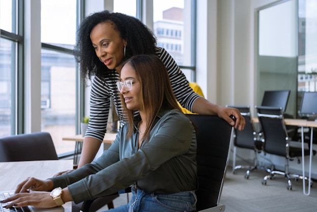 A woman stands next to a young woman who is seated at a table in front of a laptop and explains how to complete a task.