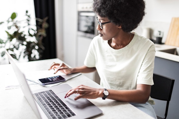  A woman entrepreneur works from home at her kitchen table calculating payments using her smartphone and then entering them into her laptop computer.