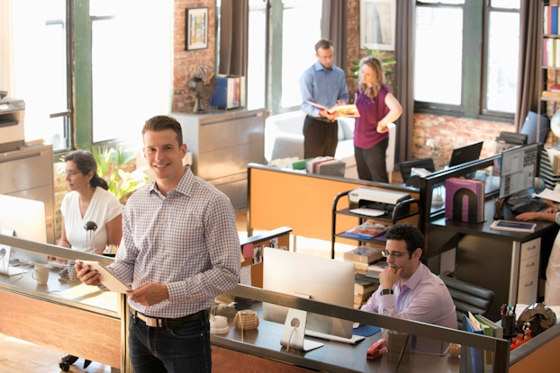 An elevated shot of a busy, modern office is shown. Several people are working on their computers or are talking while looking at a file. A man standing in the foreground smiles at the viewer.